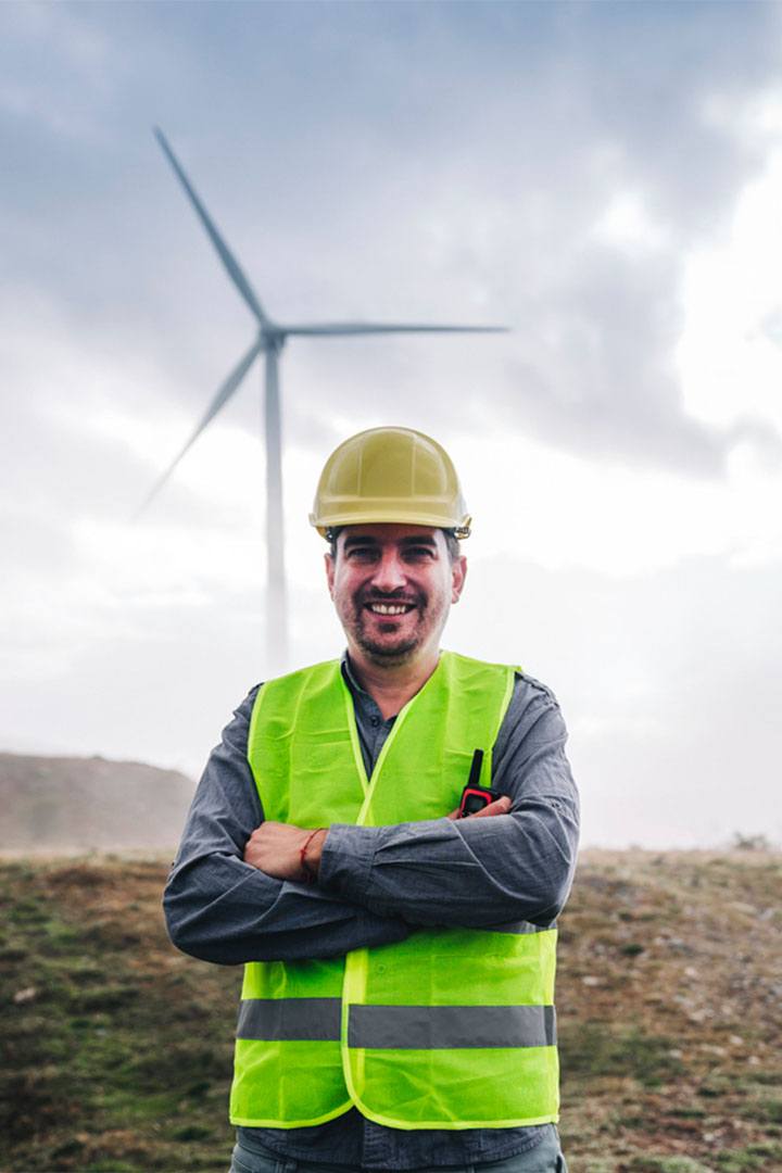 Young male in high visibility jacket and PPE standing in wind farm in engineer overalls