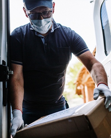 Delivery driver wearing a face mask and white gloves lifting a large cardboard box from the inside of a van through side hatch.