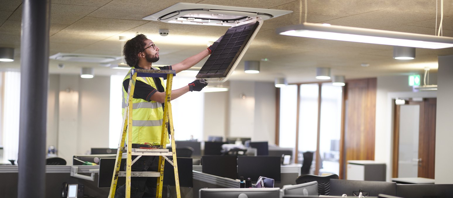 Lone working young male service engineer, inspecting air conditioning unit in empty office space.