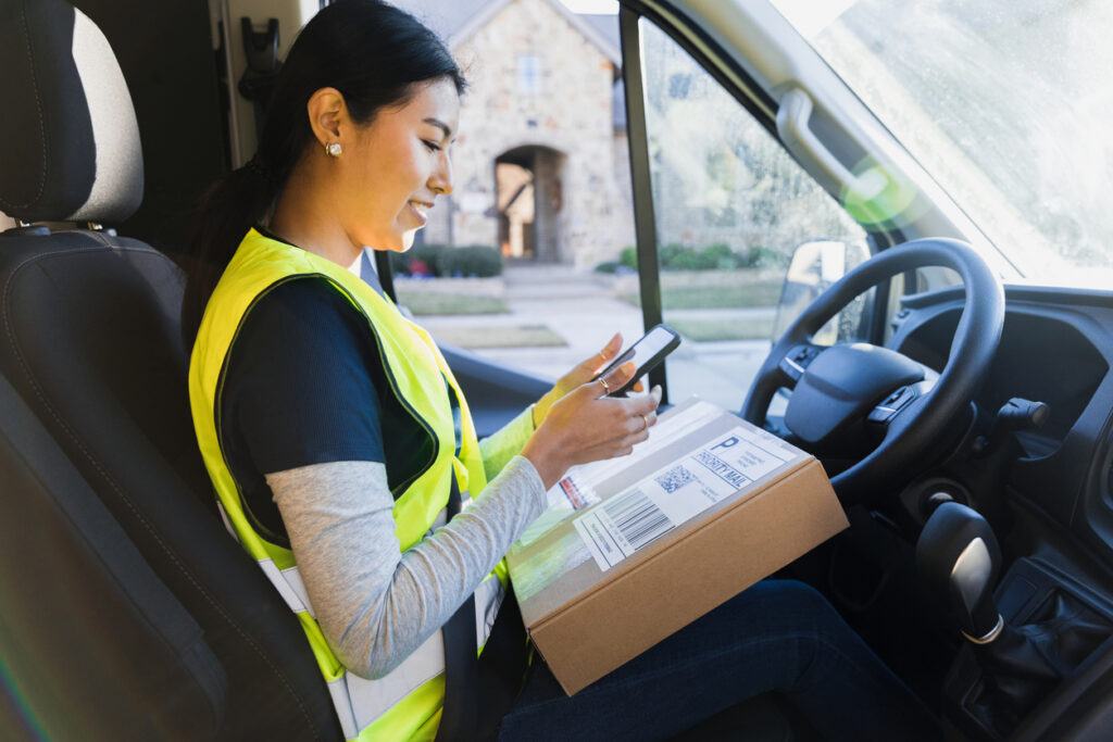 Female delivery driver inspects lone working device before delivering package to unseen customer.