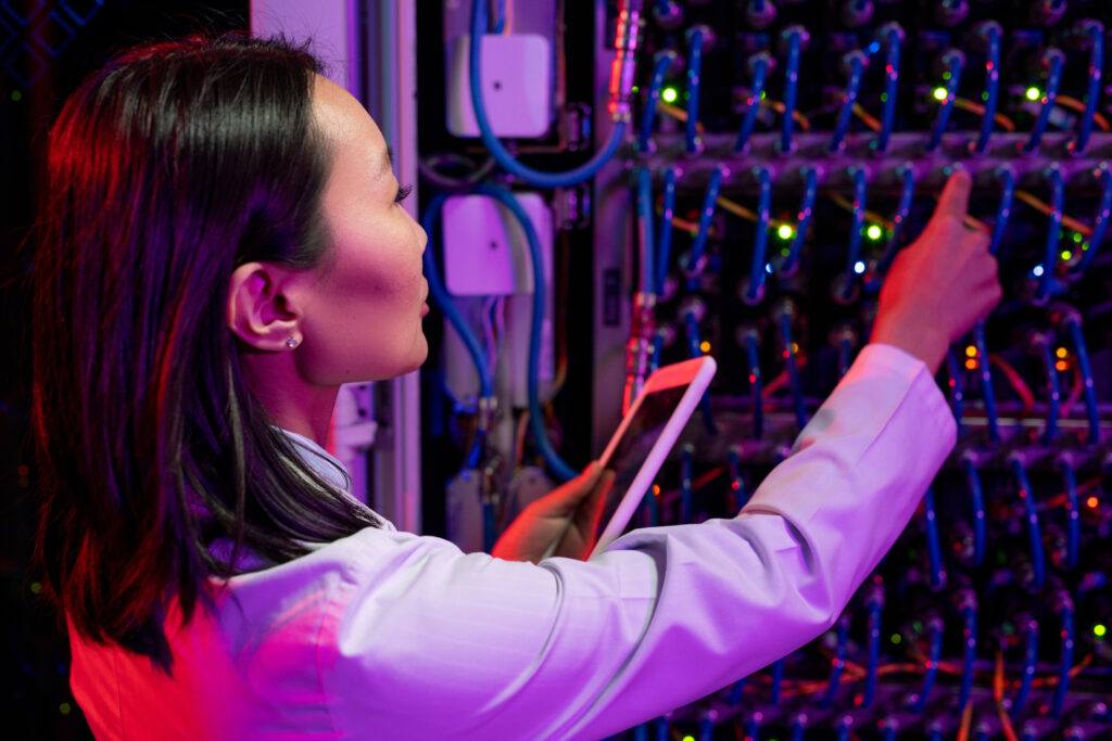 Rear view of busy Asian female network engineer in lab coat connecting cables in switches using tablet in dark room